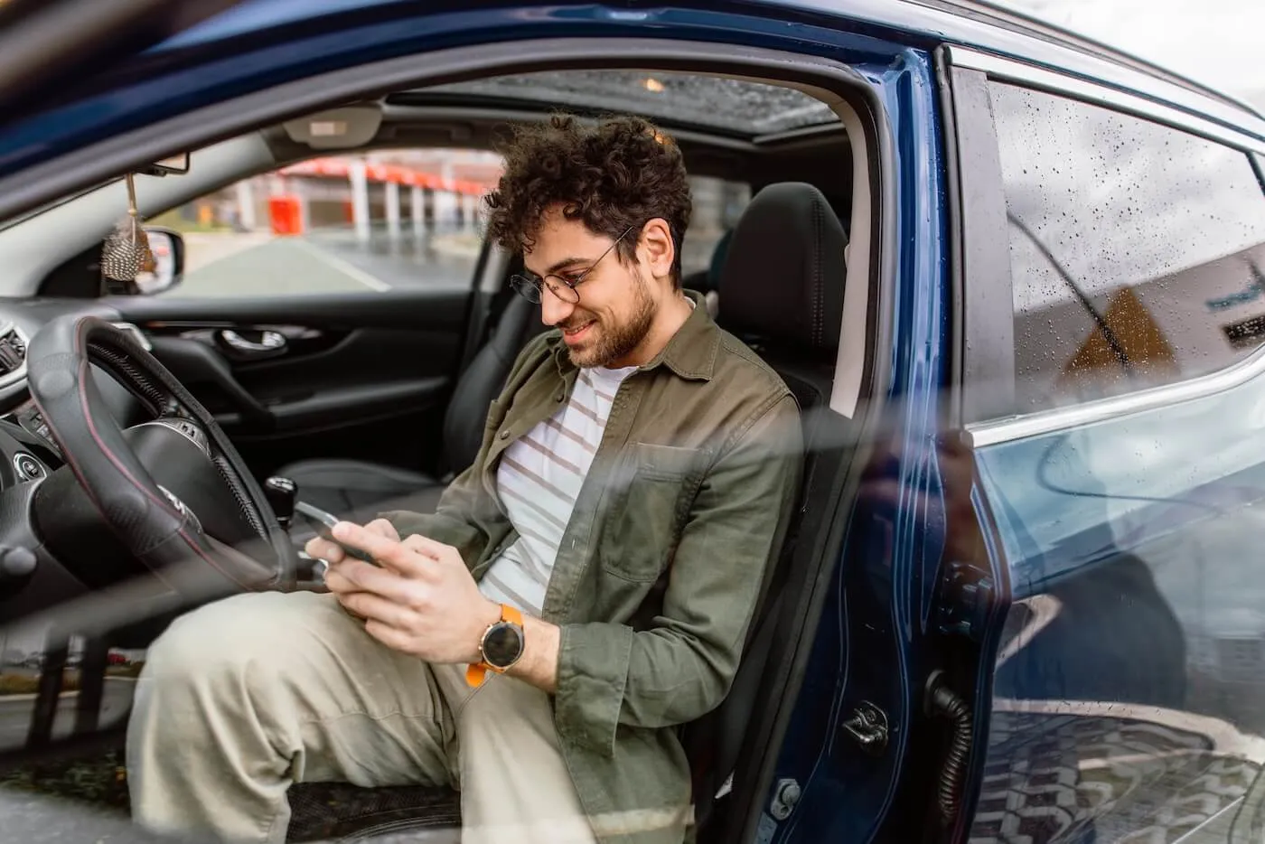 Smiling young man using his phone while sitting in a parked blue car with an open door