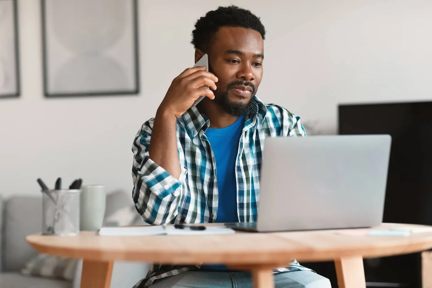 Focused young man sitting at a home desk, talking on a smartphone while working on a laptop, with notebooks and office items on a round wooden table in a modern interior.