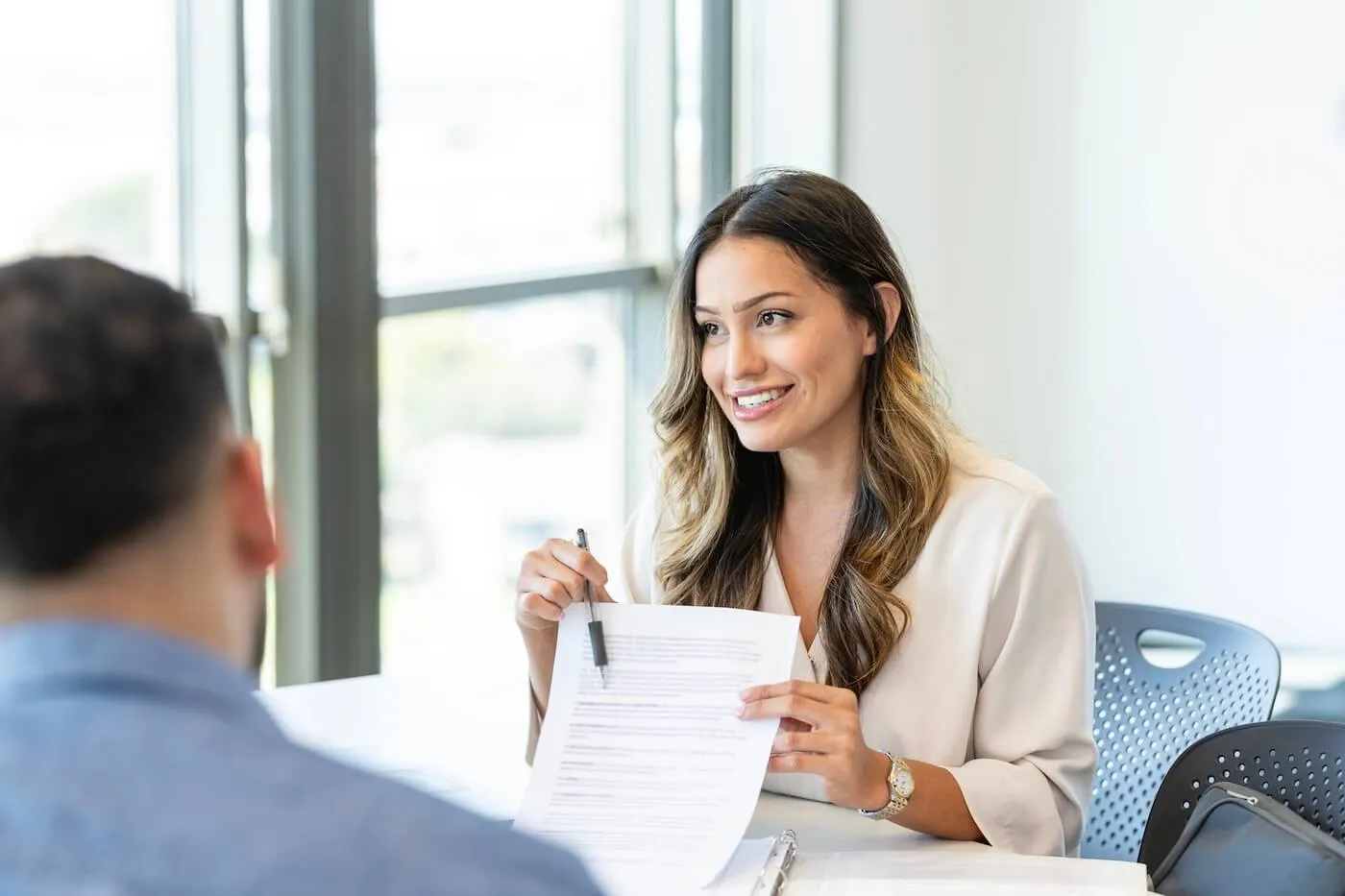 Female agent seated across from a client in a modern office, holding a document and pointing to information with a pen during a consultation. A binder and paperwork are visible on the table, with large windows and contemporary office seating in the background.