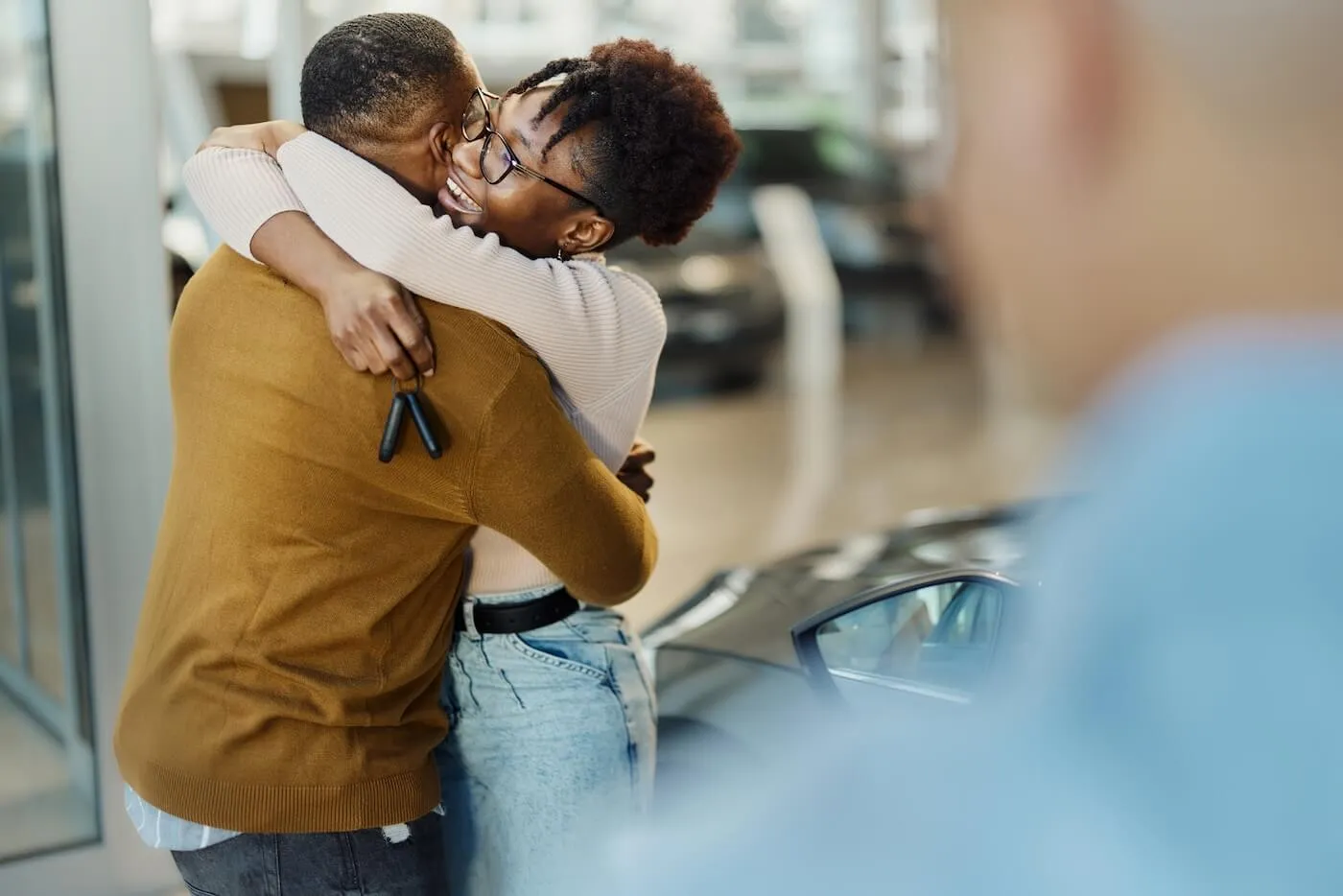 Happy couple embraces in the car dealership, with the woman holding car keys and the sales person standing nearby