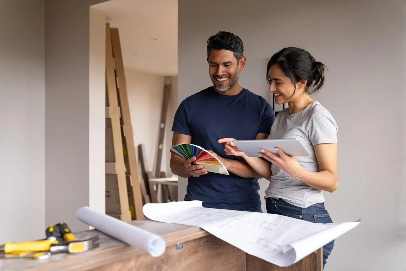 Happy couple standing at a worktable indoors, reviewing a tablet and a fan of color samples, with rolled and spread architectural plans on the table and construction materials stacked in the background.
