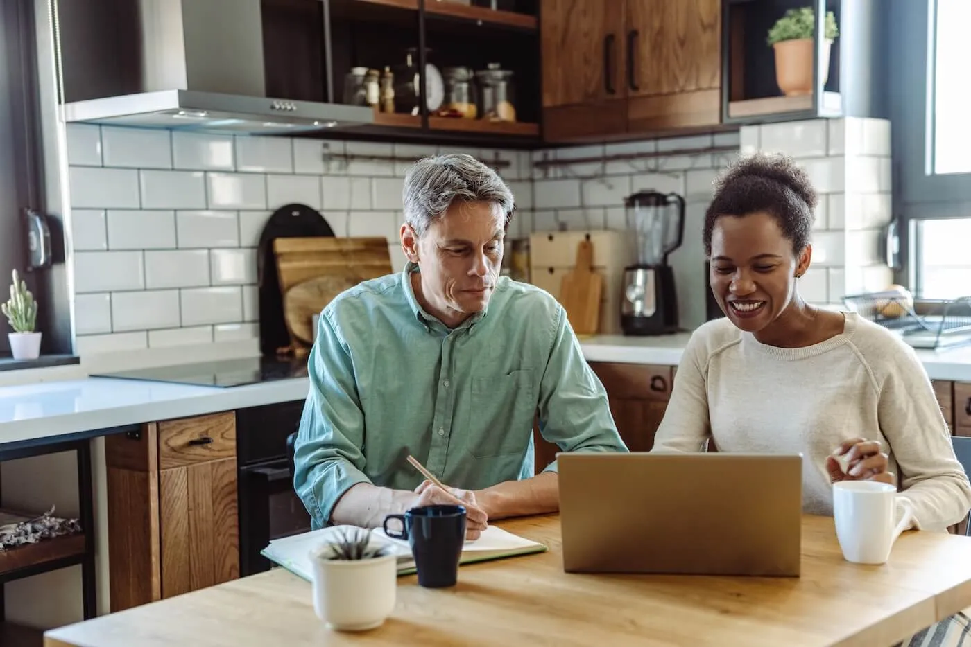 Mature couple using laptop in the kitchen while making notes, with the mugs standing on the table