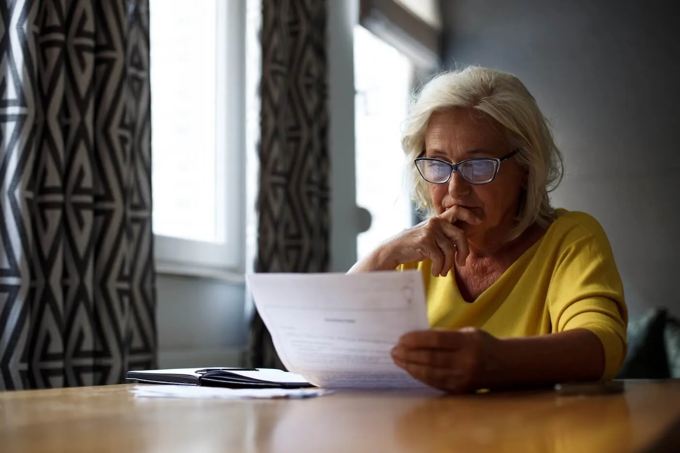 Focused senior woman wearing glasses seated at a table near a window, reviewing a printed document with a notebook nearby in a home setting.