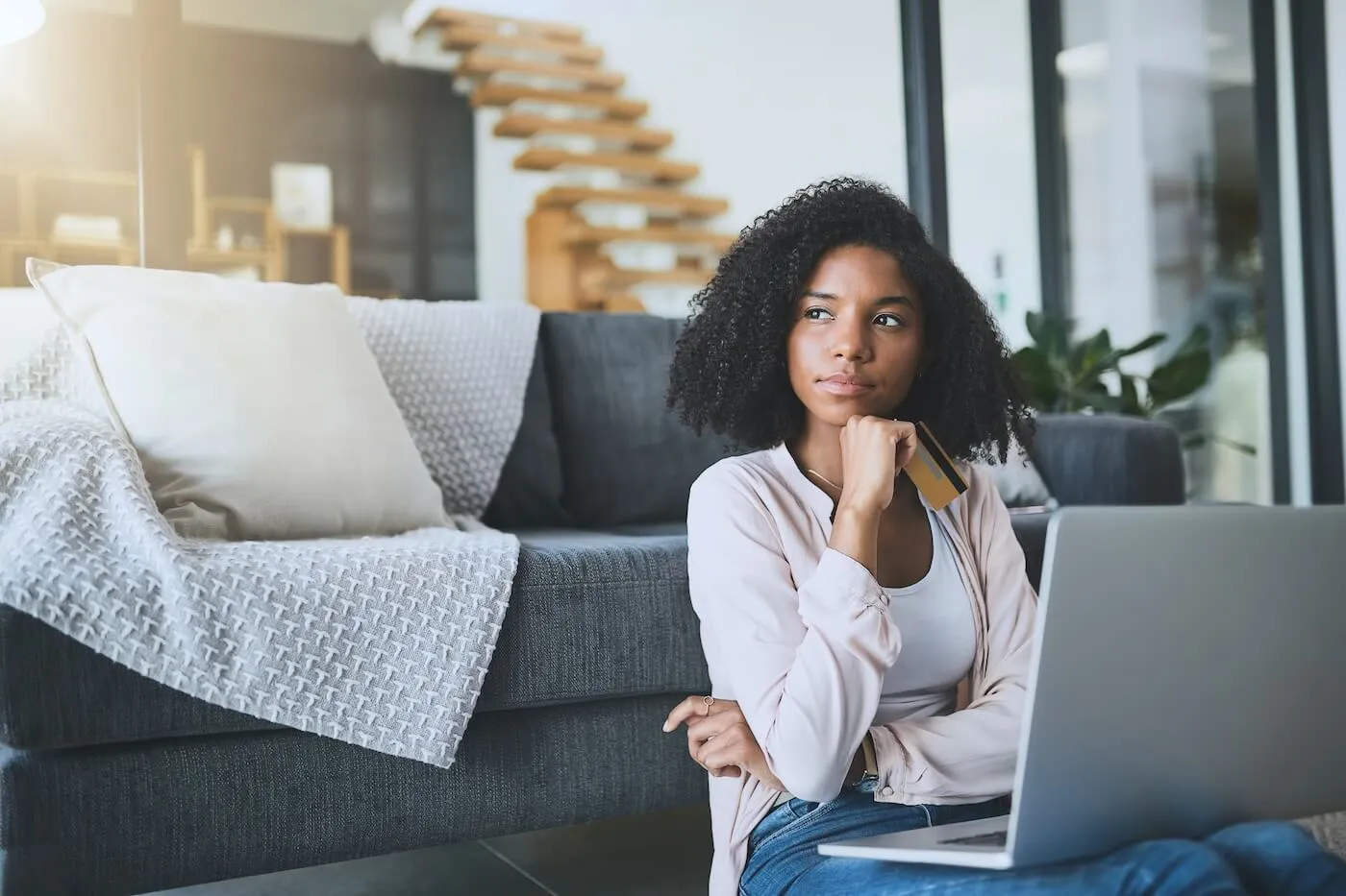Pensive woman sitting on the floor next to a couch in the living room with a laptop and holding a credit card