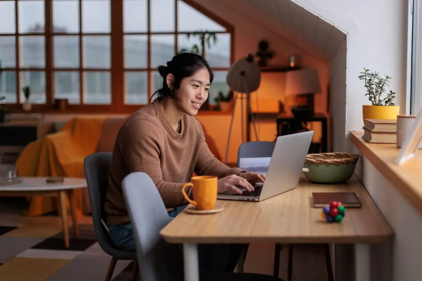 Smiling young woman sits at a wooden desk using a laptop in a cozy, softly lit room, with a mug, books, and small decor items on the table and a living area in the background.