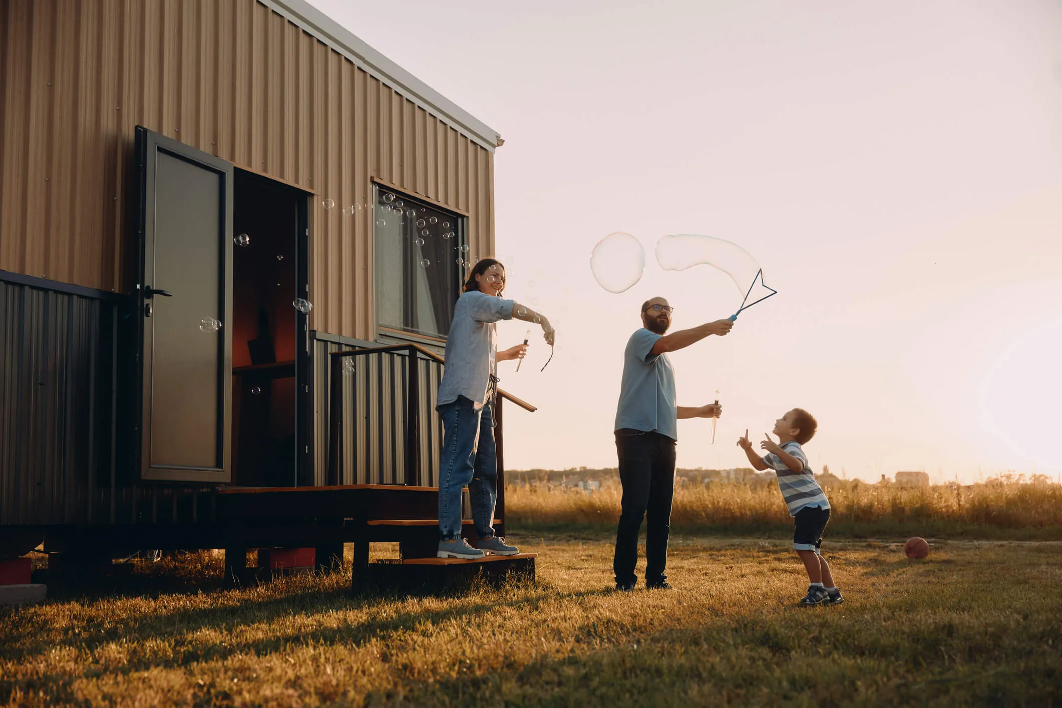 Happy family of three outdoors beside a small manufactured house, with two adults using bubble wands while a child reaches toward floating soap bubbles on a grassy yard at sunset.
