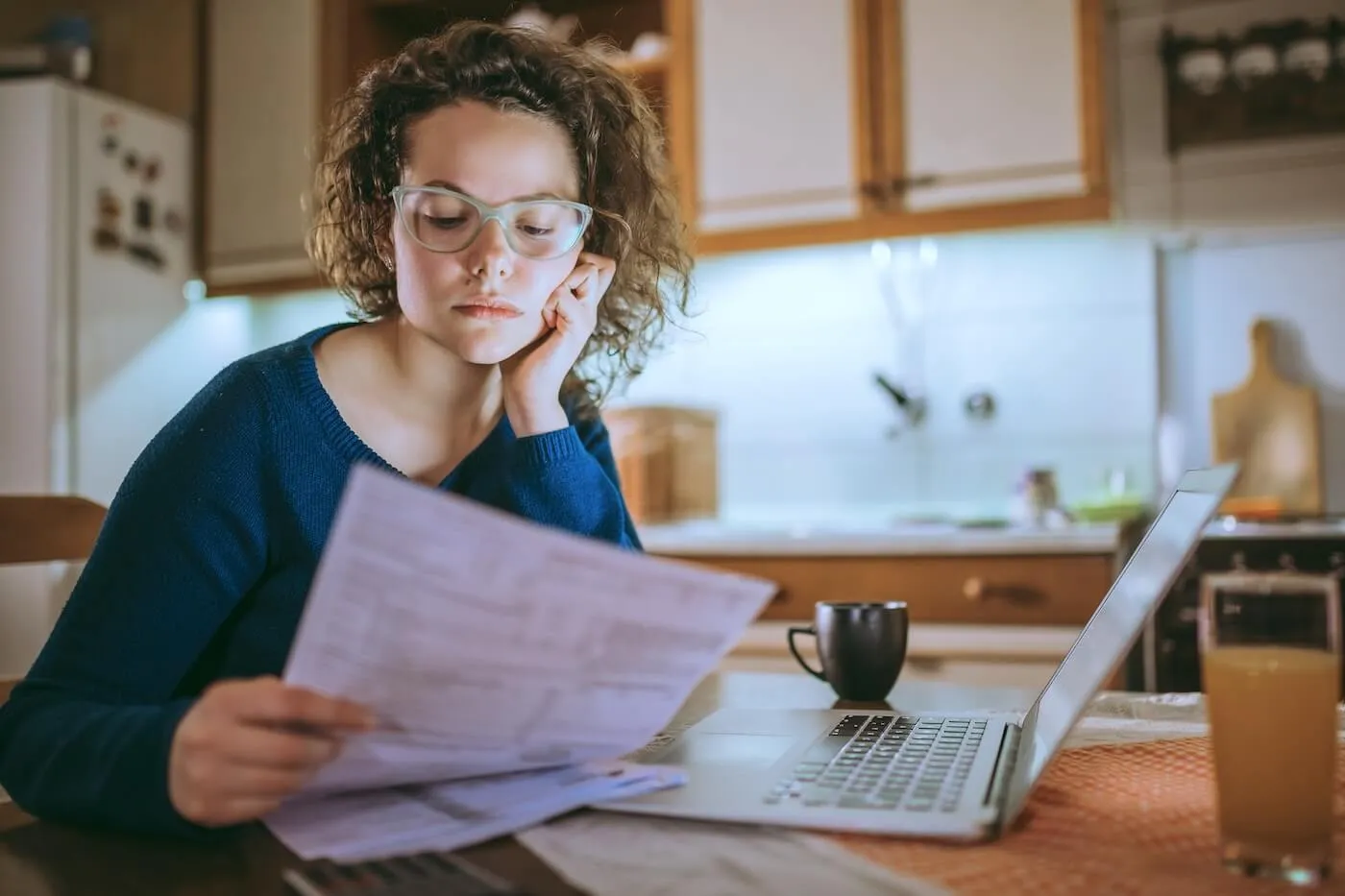 Pensive woman seated at a kitchen table reading papers, with an open laptop, a coffee mug, and a glass of orange juice nearby, wooden cabinets and kitchen items visible in the background.