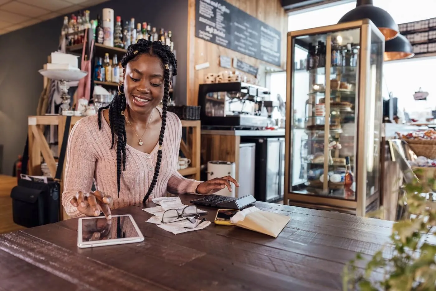 Small business owner seated at a café counter reviewing receipts and financial paperwork while using a tablet. A calculator, smartphone, eyeglasses, and business documents are spread across the table, with coffee shop equipment and display cases visible in the background.