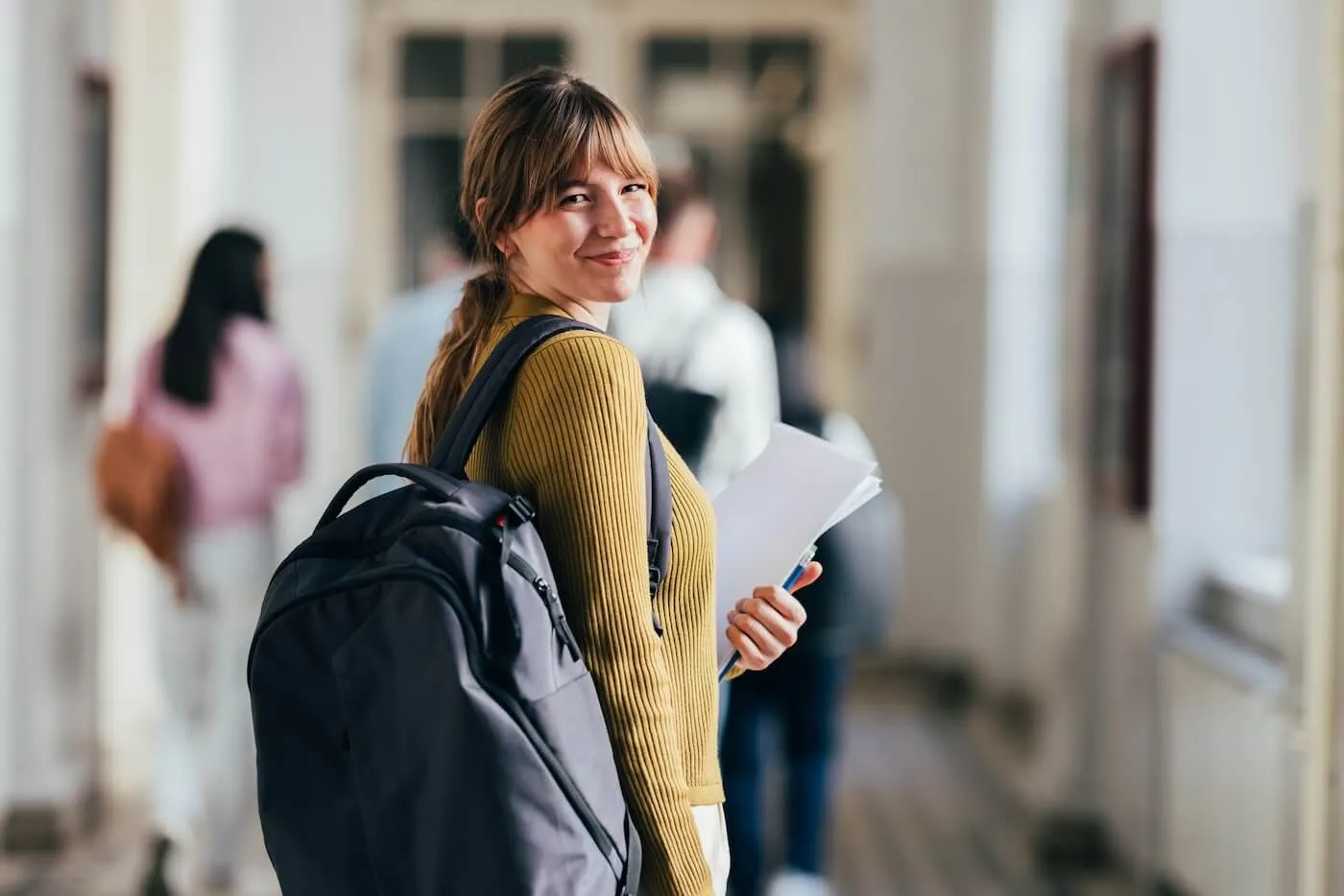 Smiling female student with a backpack holding notebooks while walking through a campus hallway.