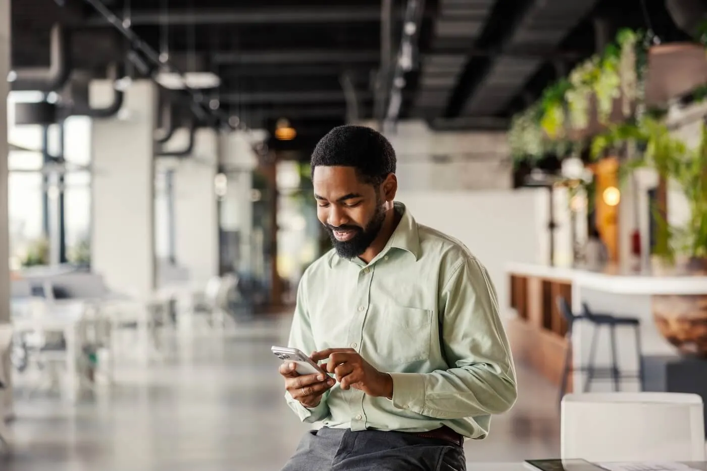 Smiling man seated in a modern office lobby or coworking space using a smartphone. The open workspace features large windows, indoor plants, tables, and contemporary furnishings, illustrating mobile banking, digital communication, or business productivity on the go.