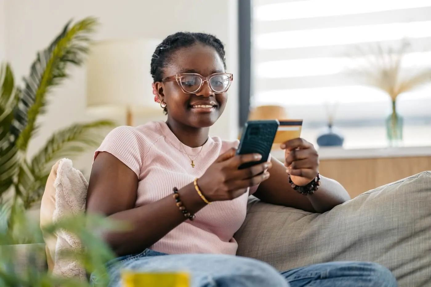 Smiling young woman seated on a sofa in a bright living room, holding a smartphone in one hand and a credit card in the other. Houseplants, a large window, and comfortable home furnishings are visible in the background.