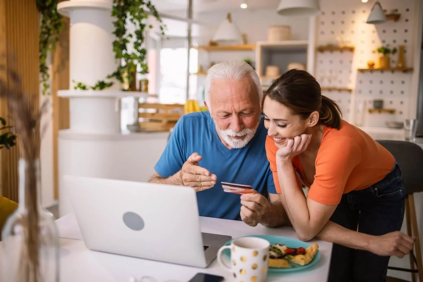 Older man and younger woman sitting at a kitchen table, using a laptop and holding a credit card while discussing an online payment.