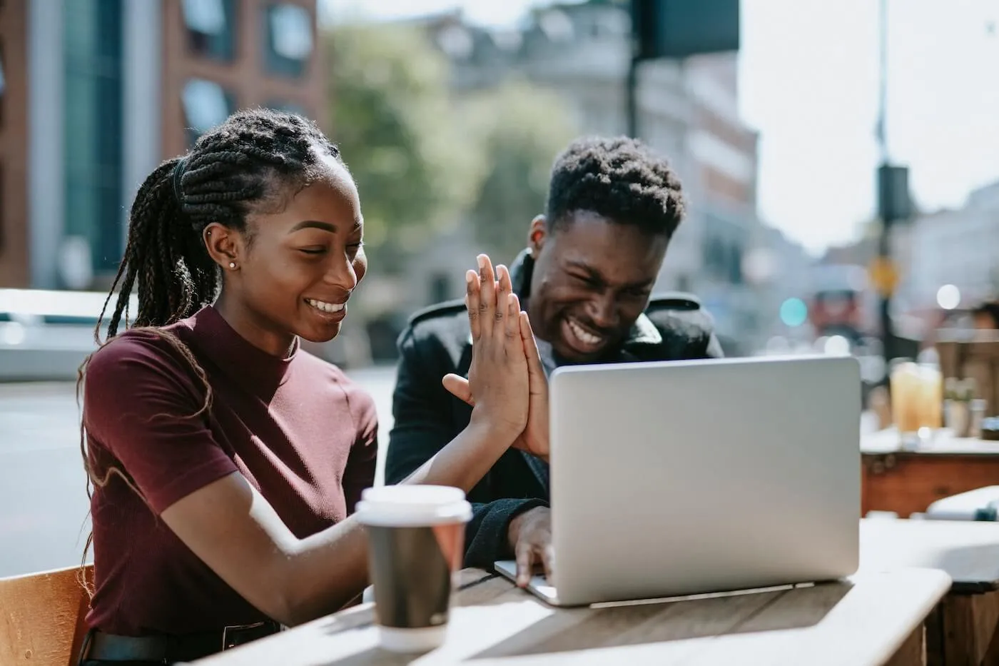 Happy couple sits at an outdoor table using a laptop and high-five, with takeaway coffee cups and a city street setting in the background.