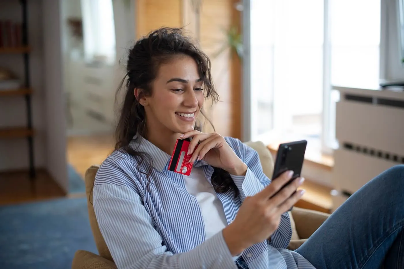 Smiling young woman sitting in a chair while using a smartphone and holding a credit card