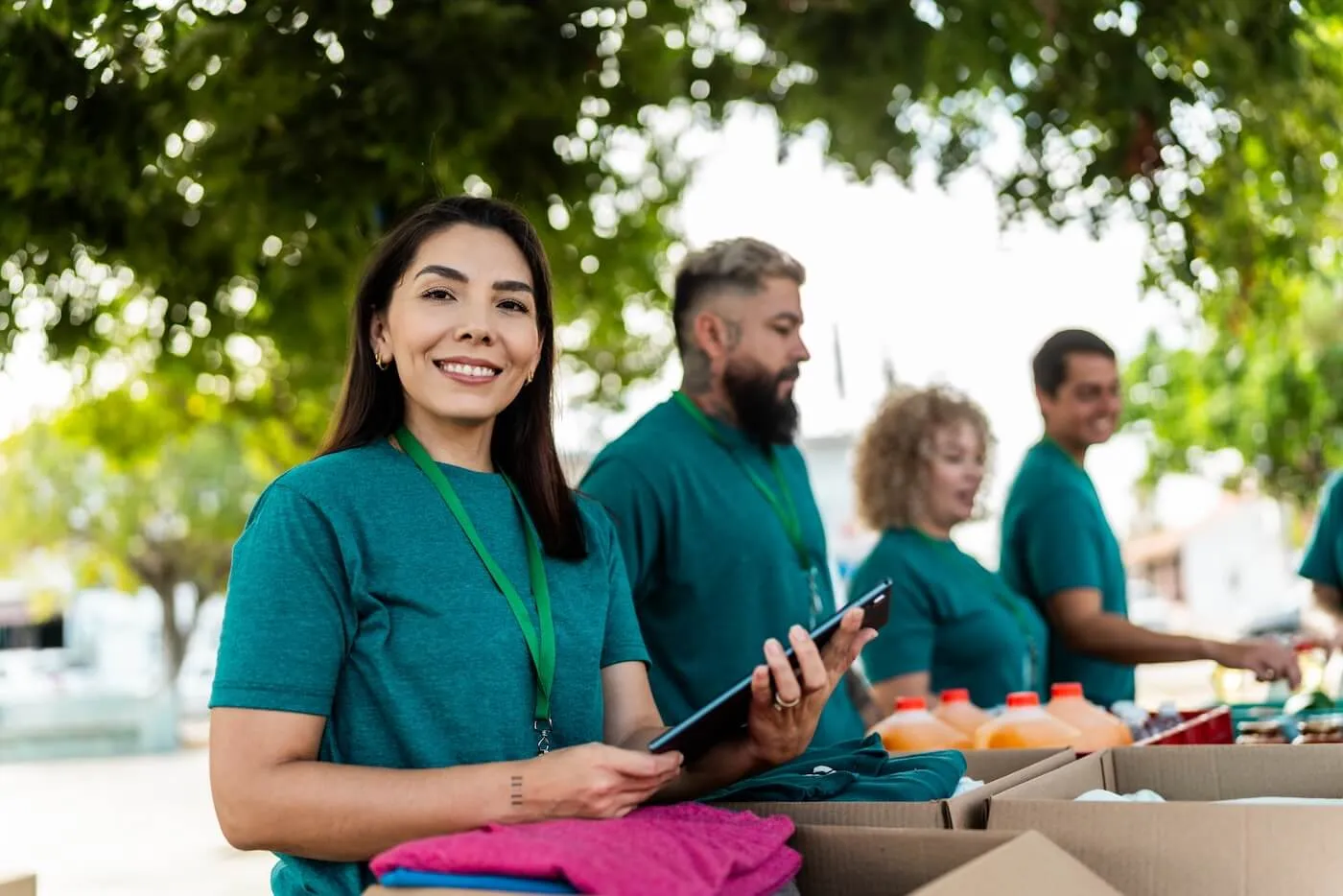 Group of volunteers in green T‑shirts distributing goods outdoors, with a smiling woman holding a tablet and looking at the camera