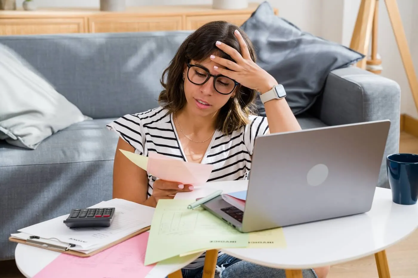 Tired woman seated at a coffee table reviewing financial documents and bills while using a laptop, with a calculator, paperwork, and a mug nearby in a living room setting.
