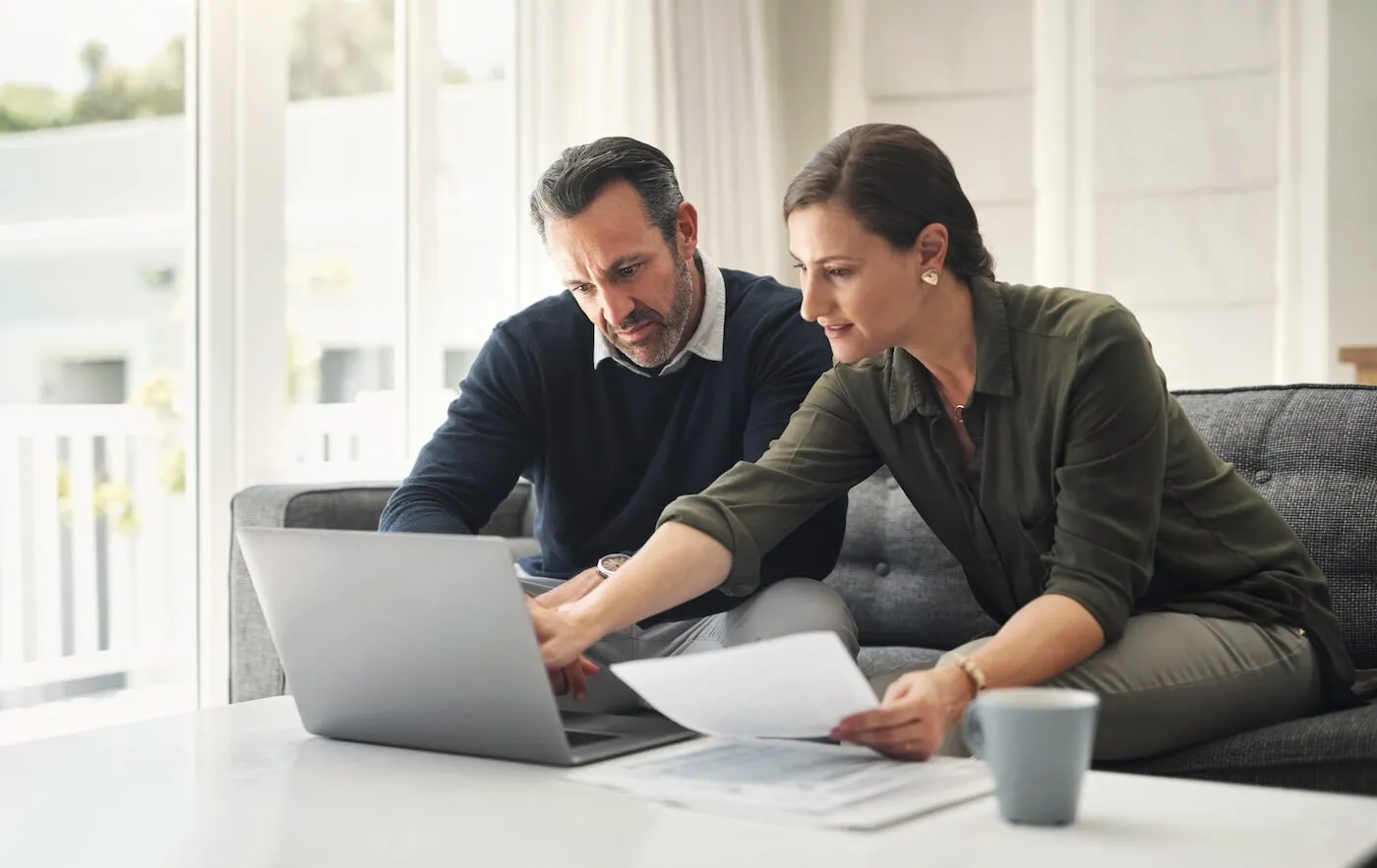 Focused couple sitting together on a sofa, looking at paperwork and a laptop placed on a table, with the woman gesturing toward the laptop screen in a bright living room.
