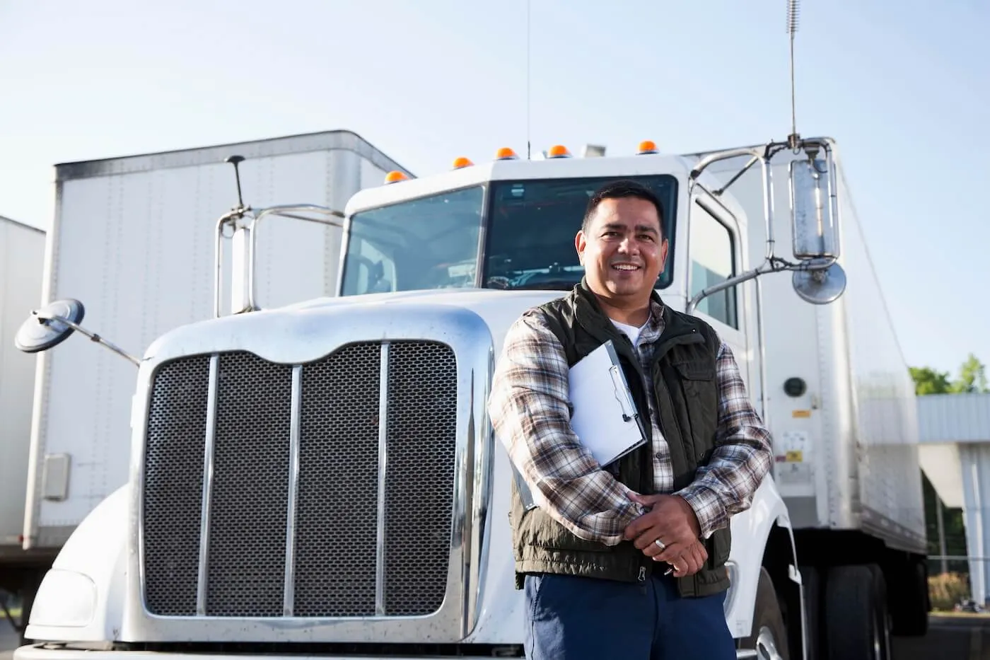 Male truck driver standing in front of a white semi-truck and trailer while holding a clipboard, with additional commercial trucks visible in a transportation or logistics yard.