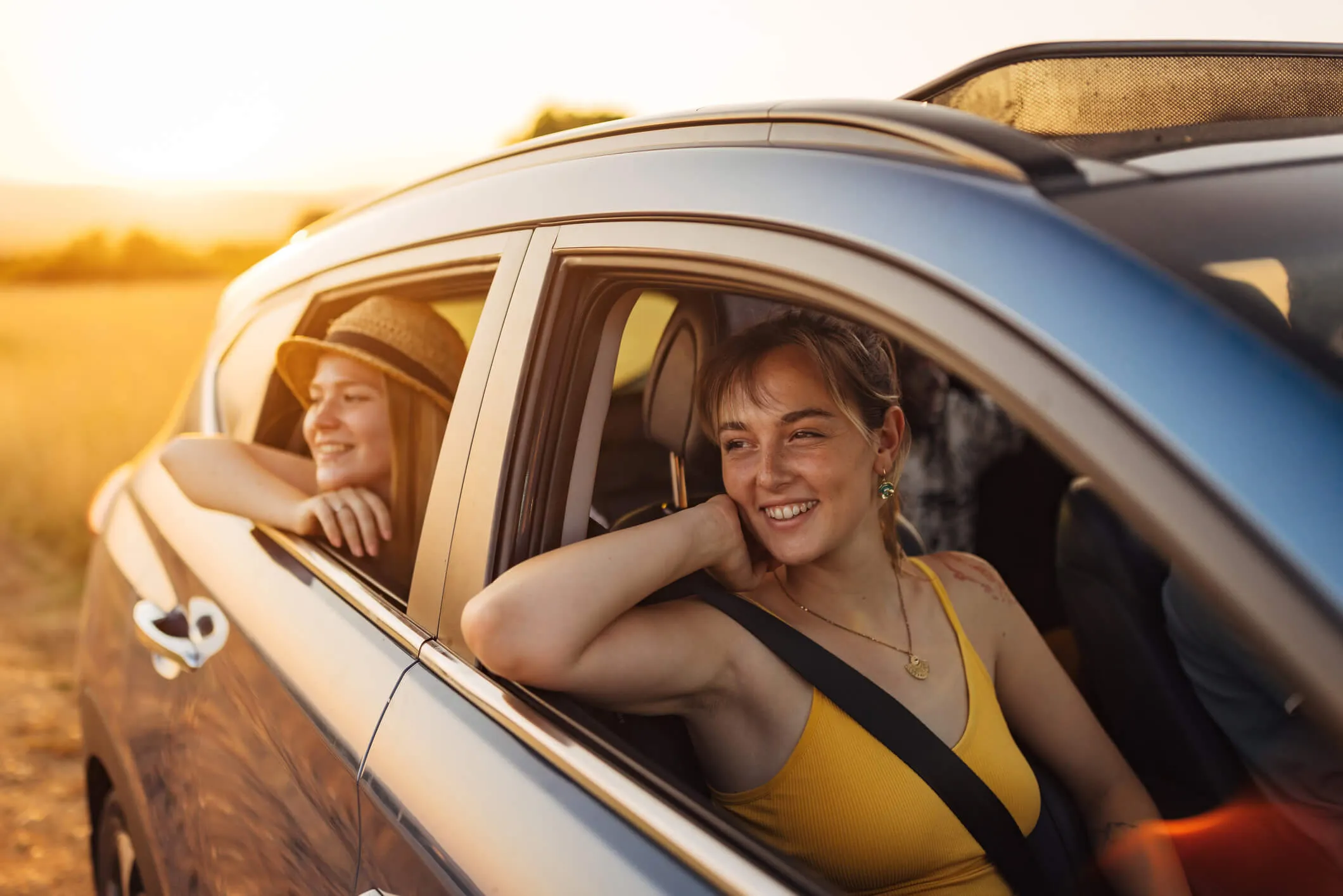 Two young females seating inside a parked car on the passenger seats with the windows open, viewed from outside the vehicle, with one person in the front seat and another seated behind, both wearing seat belts.