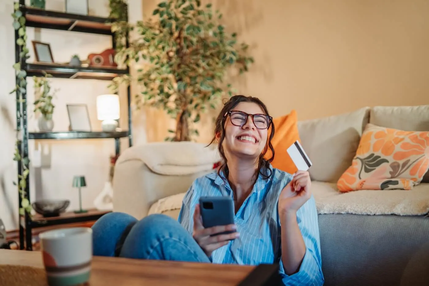Smiling young woman sitting on the floor in a living room, holding a smartphone in one hand and a credit card in the other, with a sofa, coffee table, shelving unit, and houseplants visible in the background.