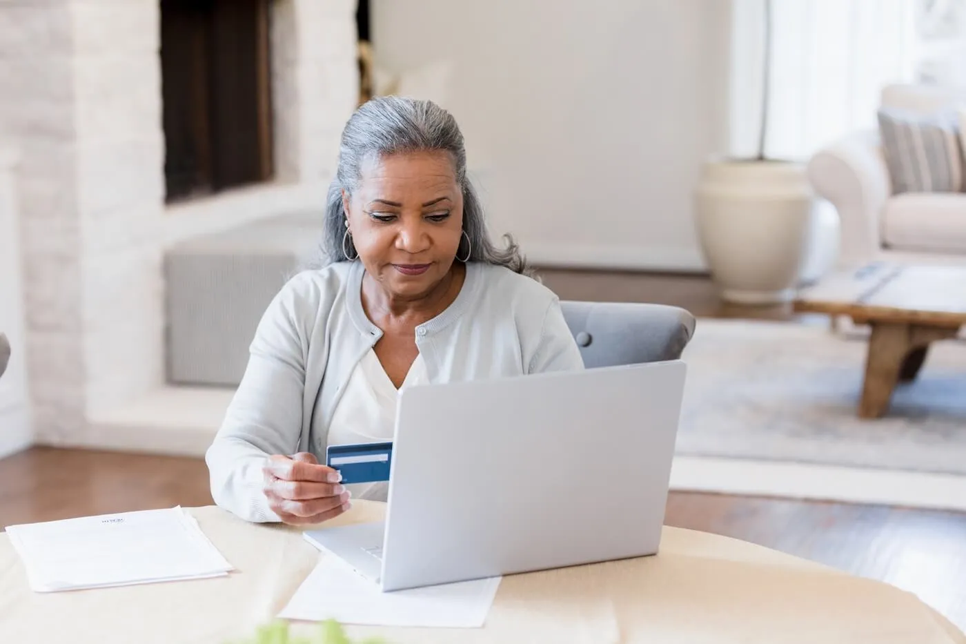 Senior woman sitting at a dining table, using a laptop while holding a credit card, with paperwork spread out in a bright living room