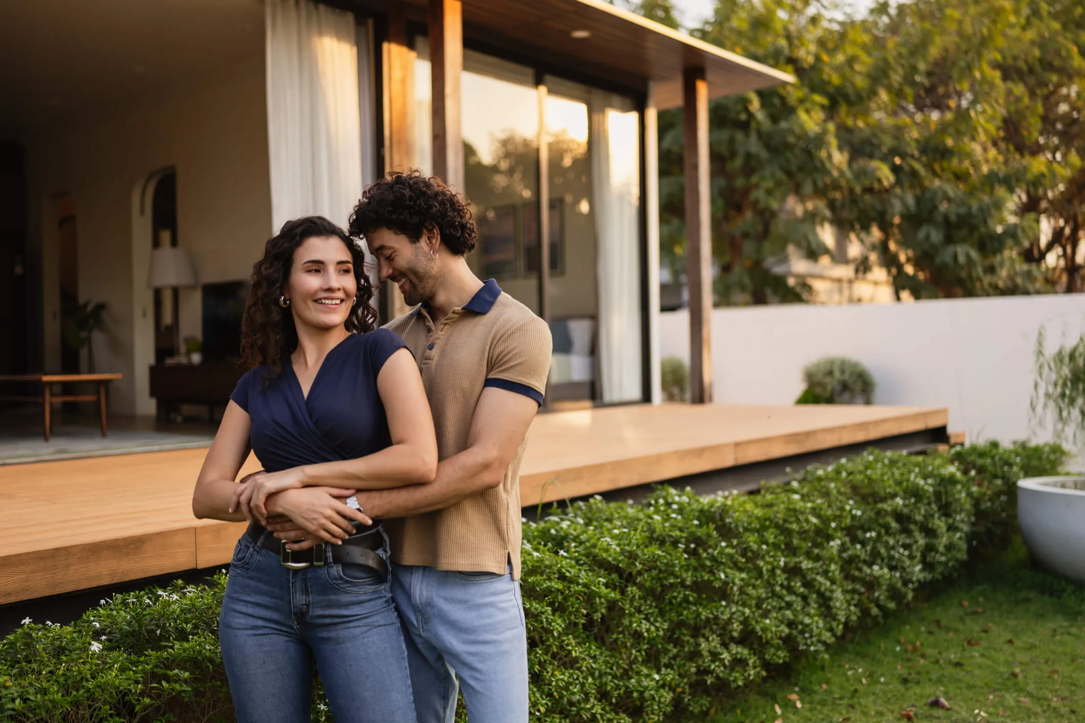 Happy couple embracing while standing in a yard outside a modern house with a wooden deck and large windows, with greenery and landscaping visible around them.