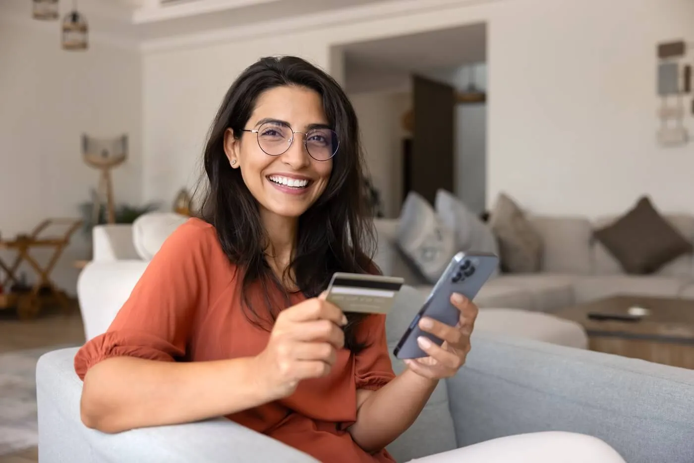 Smiling young woman using her smartphone and a credit card while sitting on a sofa in the living room