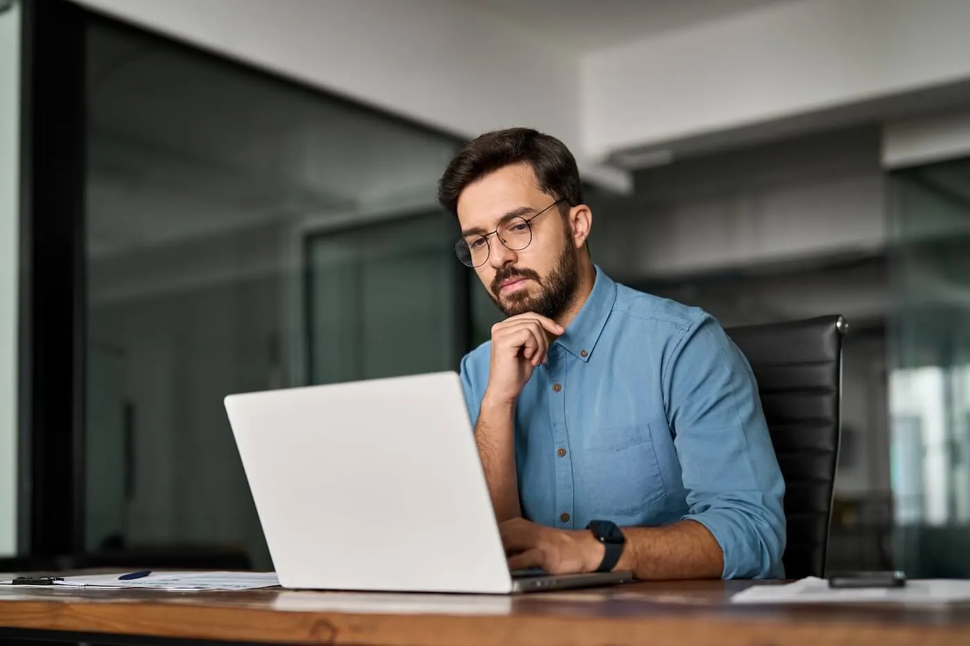 Focused young man seated at a wooden desk in a modern office setting works on an open laptop, resting one hand near the chin, with papers on the desk and glass walls and office furnishings visible in the background.