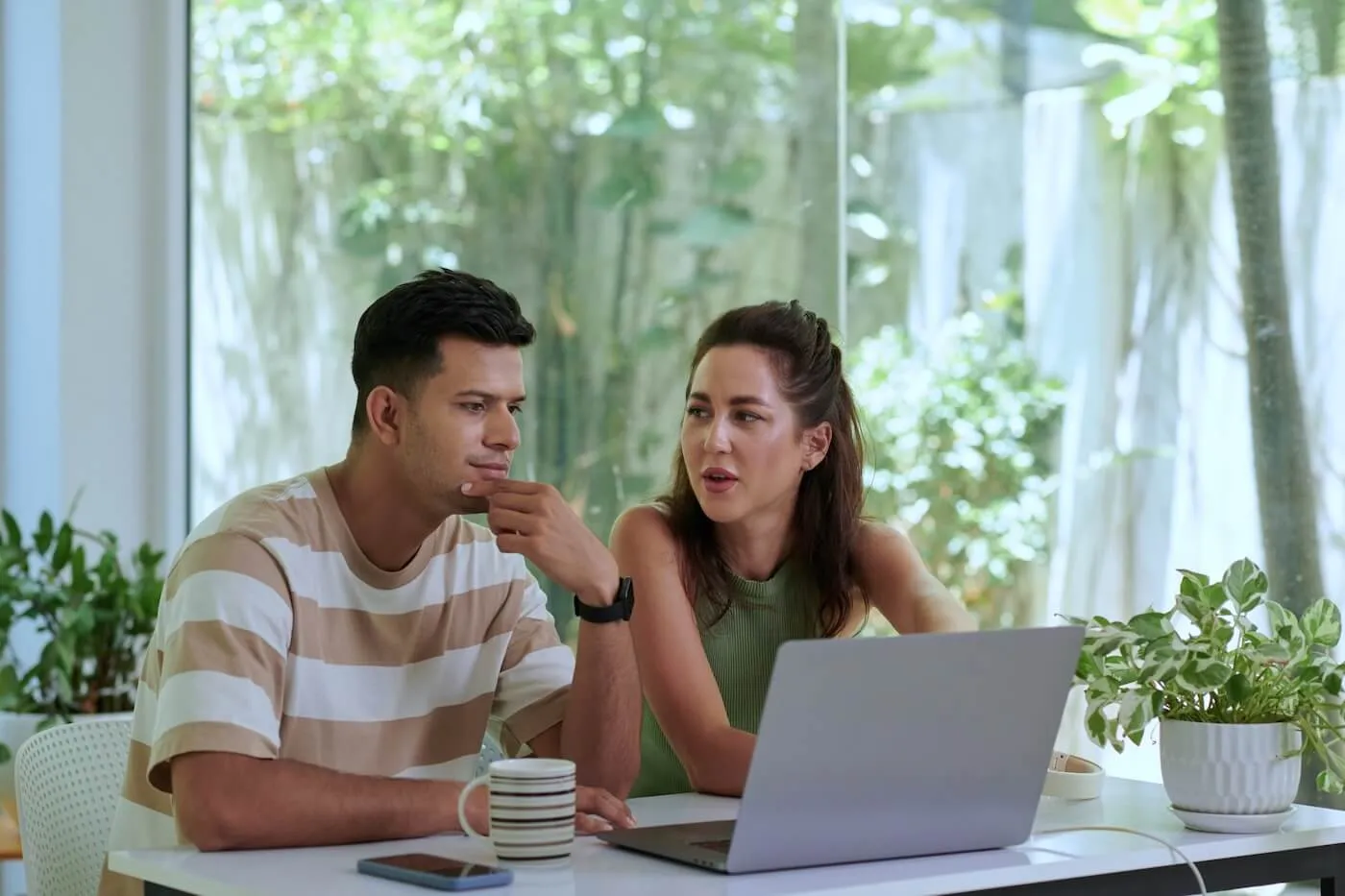 Focused couple sitting at a table indoors, looking at a laptop together, with mugs, a smartphone, and potted plants on the table and large windows in the background.