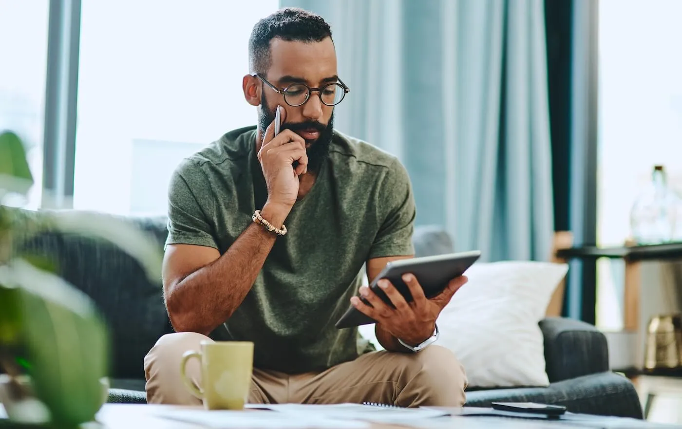 Focused man sits in a living room using a tablet while reviewing paperwork