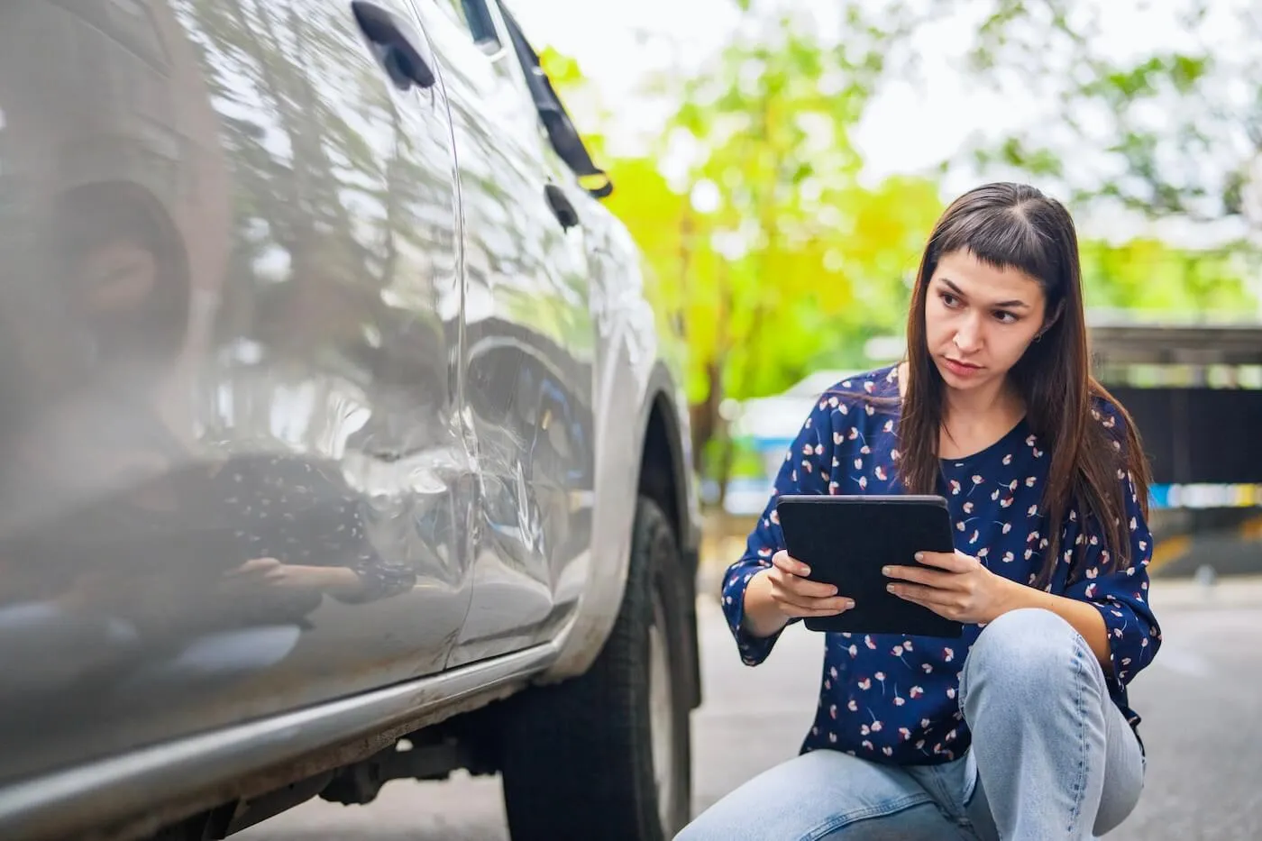Focused women kneeling beside a vehicle, using a tablet to document damage on the car’s side