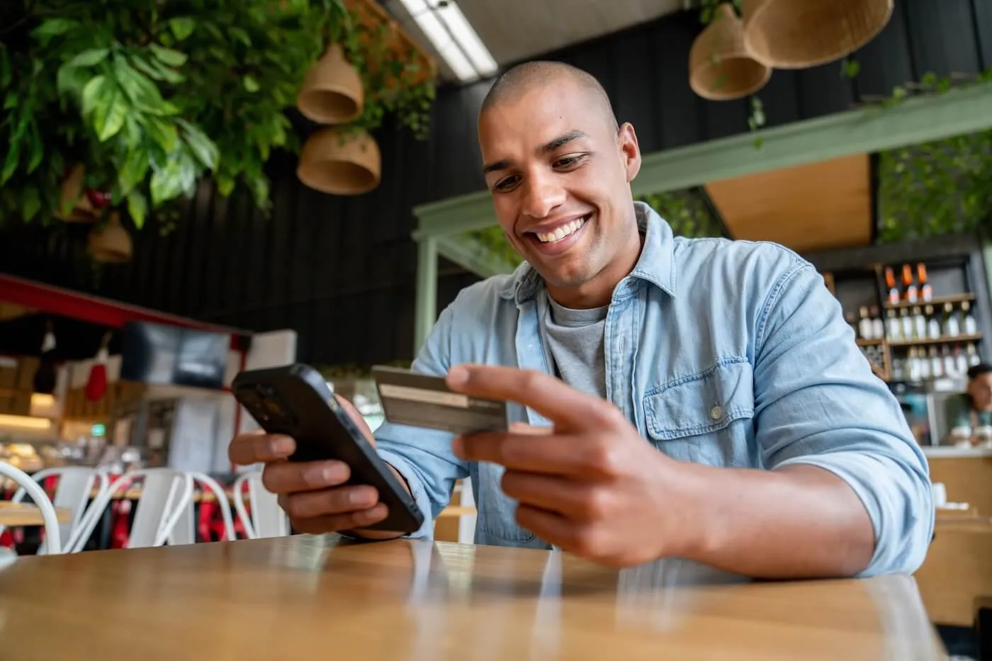 Young man person smiling while holding a credit card in one hand and using a smartphone at a café table.