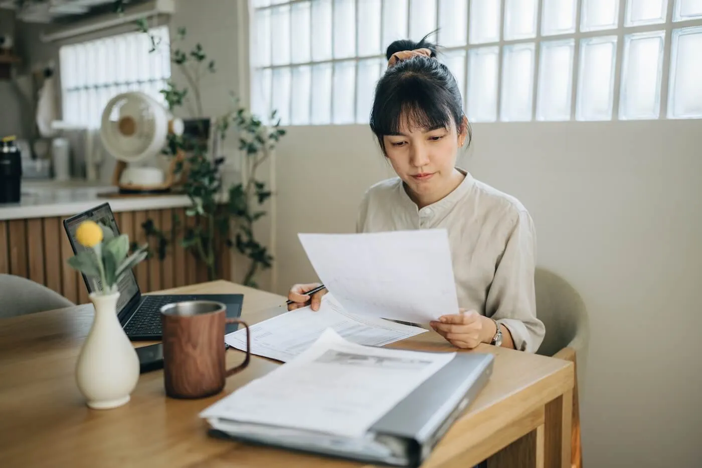 Focused woman sits at a wooden table reviewing printed documents, holding a pen and a sheet of paper, with a laptop, binder, mug, and additional papers on the table.