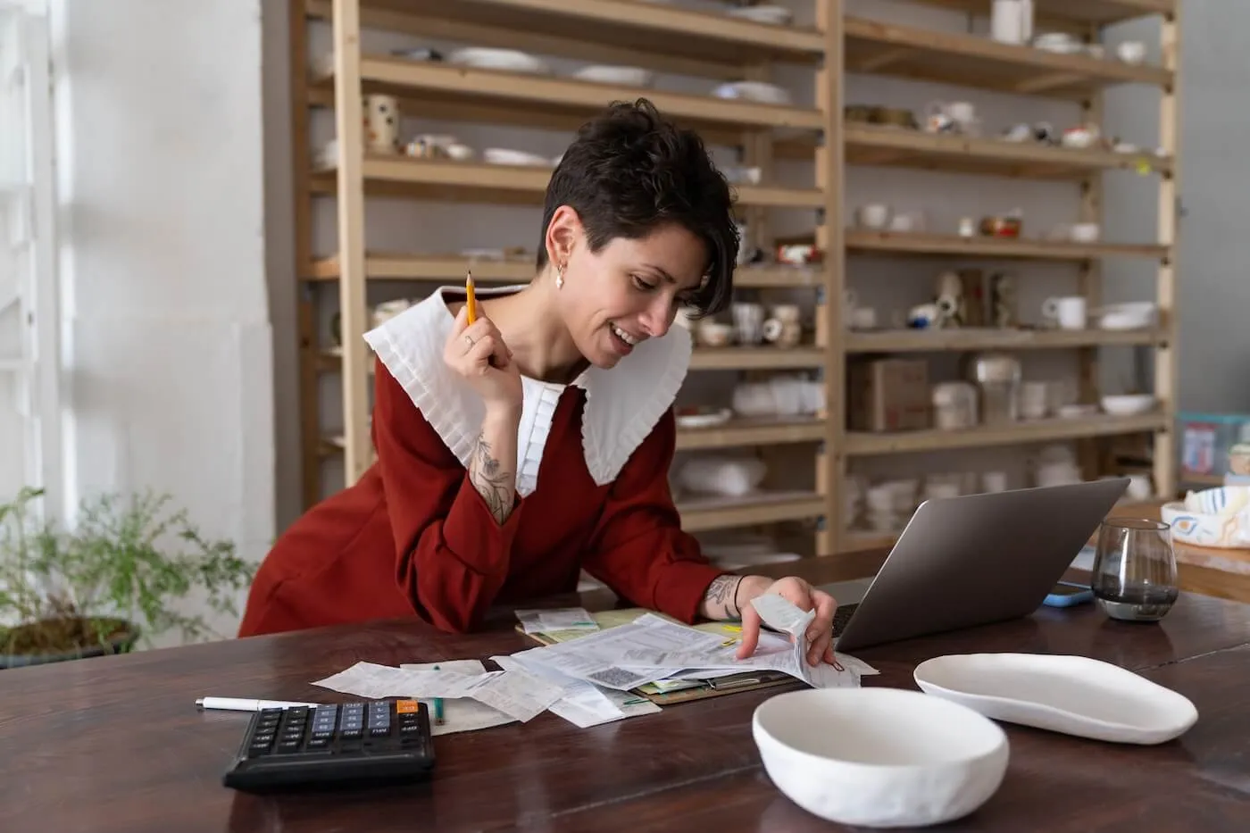 A young woman in a red dress with a white collar reviewing receipts and notes at a desk with a laptop and calculator, focused on organizing finances, with shelves of ceramic goods in the background.