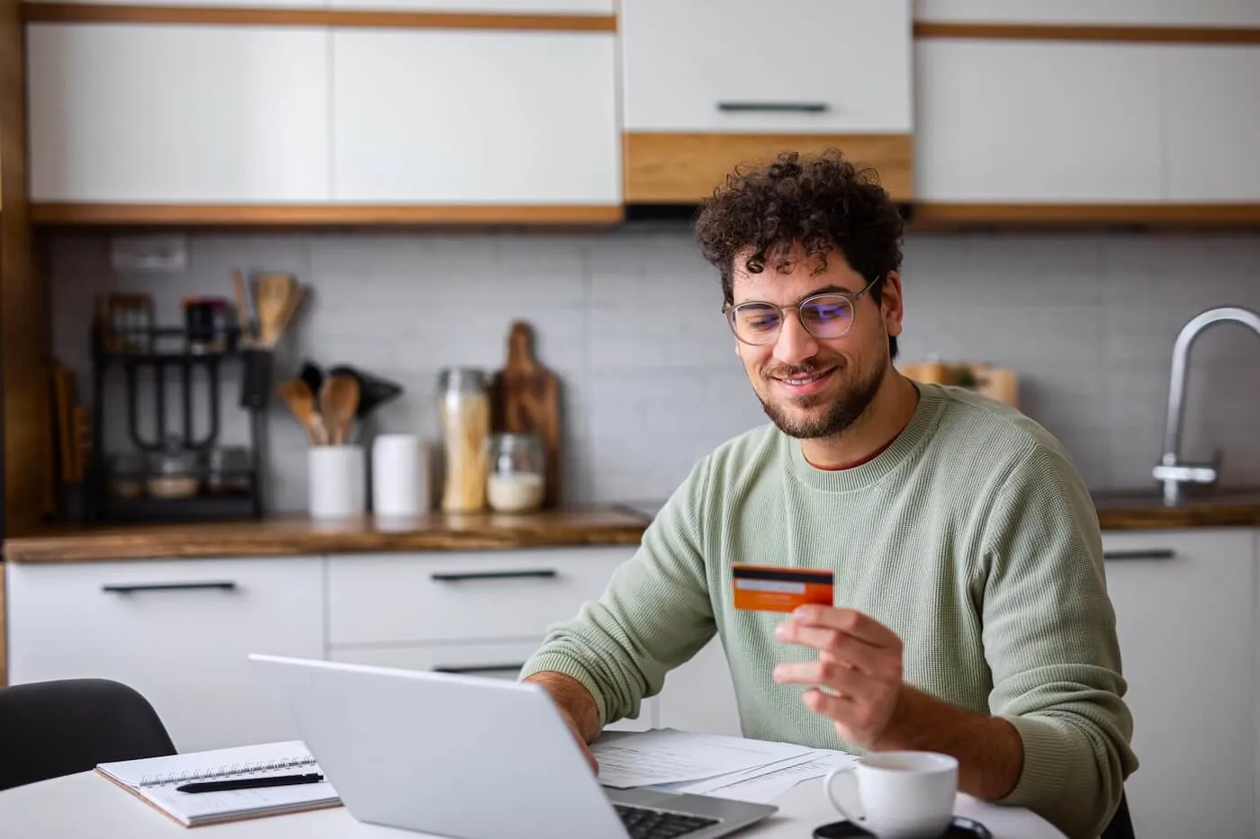 Smiling young man using his credit card online while sitting at the kitchen table with an open laptop