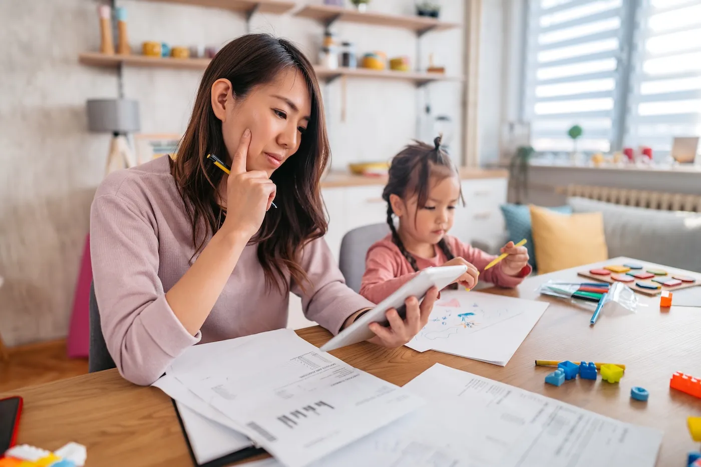 Focused young woman reviewing printed documents while a toddler girl playing beside her.