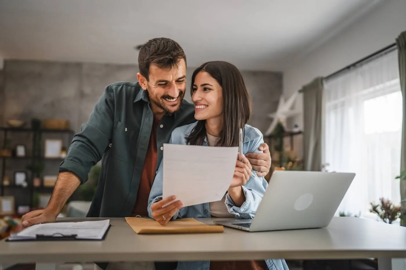 Happy couple standing close together at a table while looking at a document, with an open laptop, notebook, and papers arranged on the surface in a home setting.
