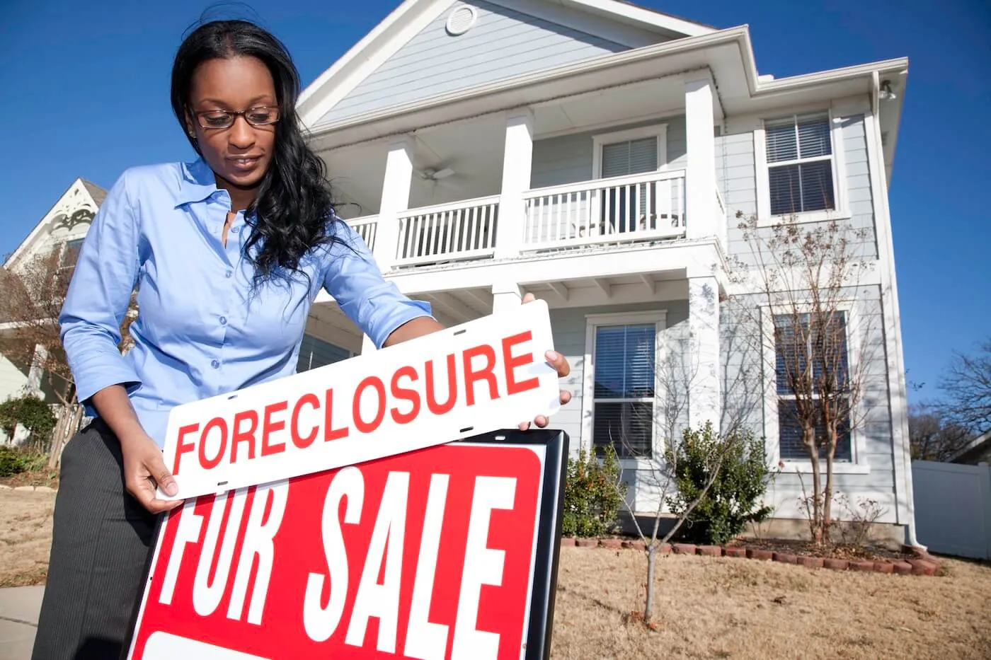 Female agent adjusting a real estate sign that reads “Foreclosure” above a “For Sale” sign, with a two‑story home visible in the background.