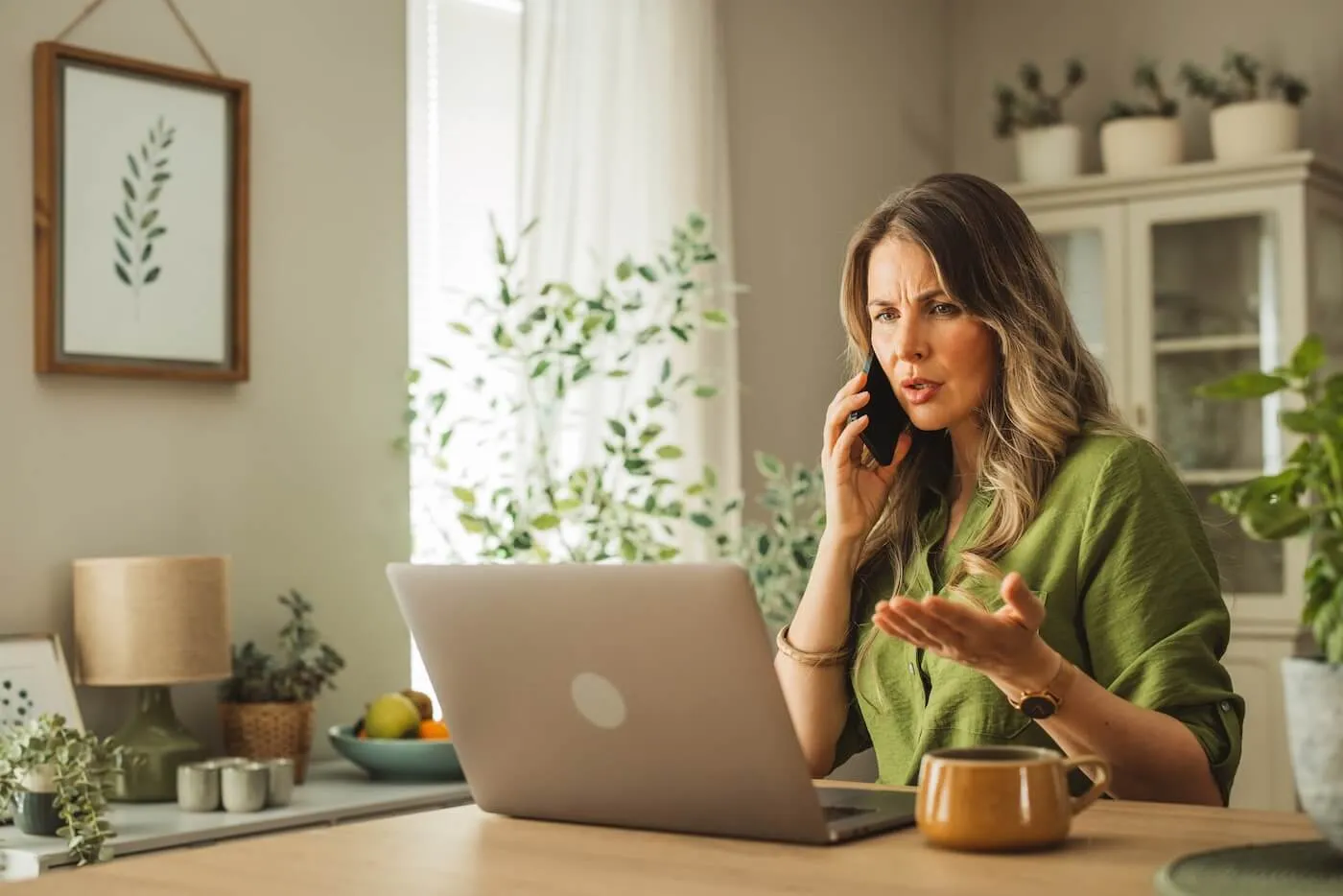 Concerned woman seated at a wooden table using a laptop while holding a smartphone to one ear and gesturing with the other hand, with a mug on the table and plants, framed artwork, and light-colored furniture visible in a bright home interior
