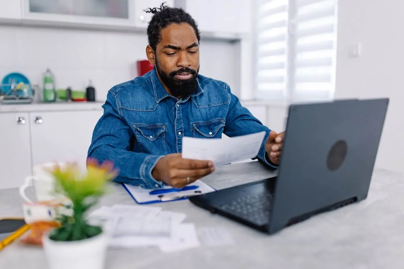 Focused man sits at a table holding papers while looking at a laptop, with additional documents and a pen on the table