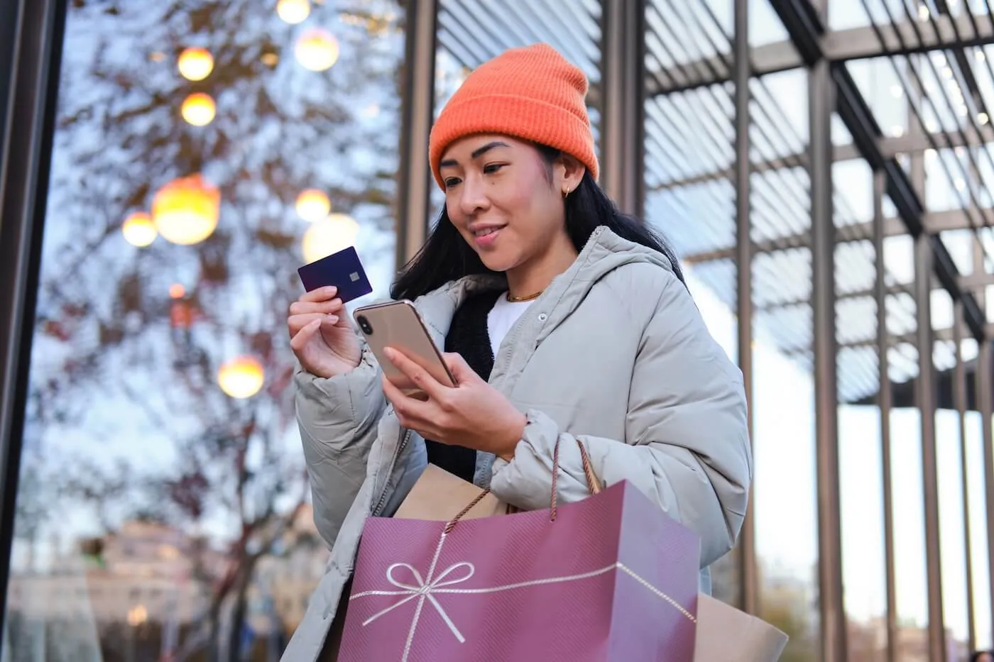 Smiling woman in winter clothes outside a shopping center, holding a festive bag, credit card, and smartphone.