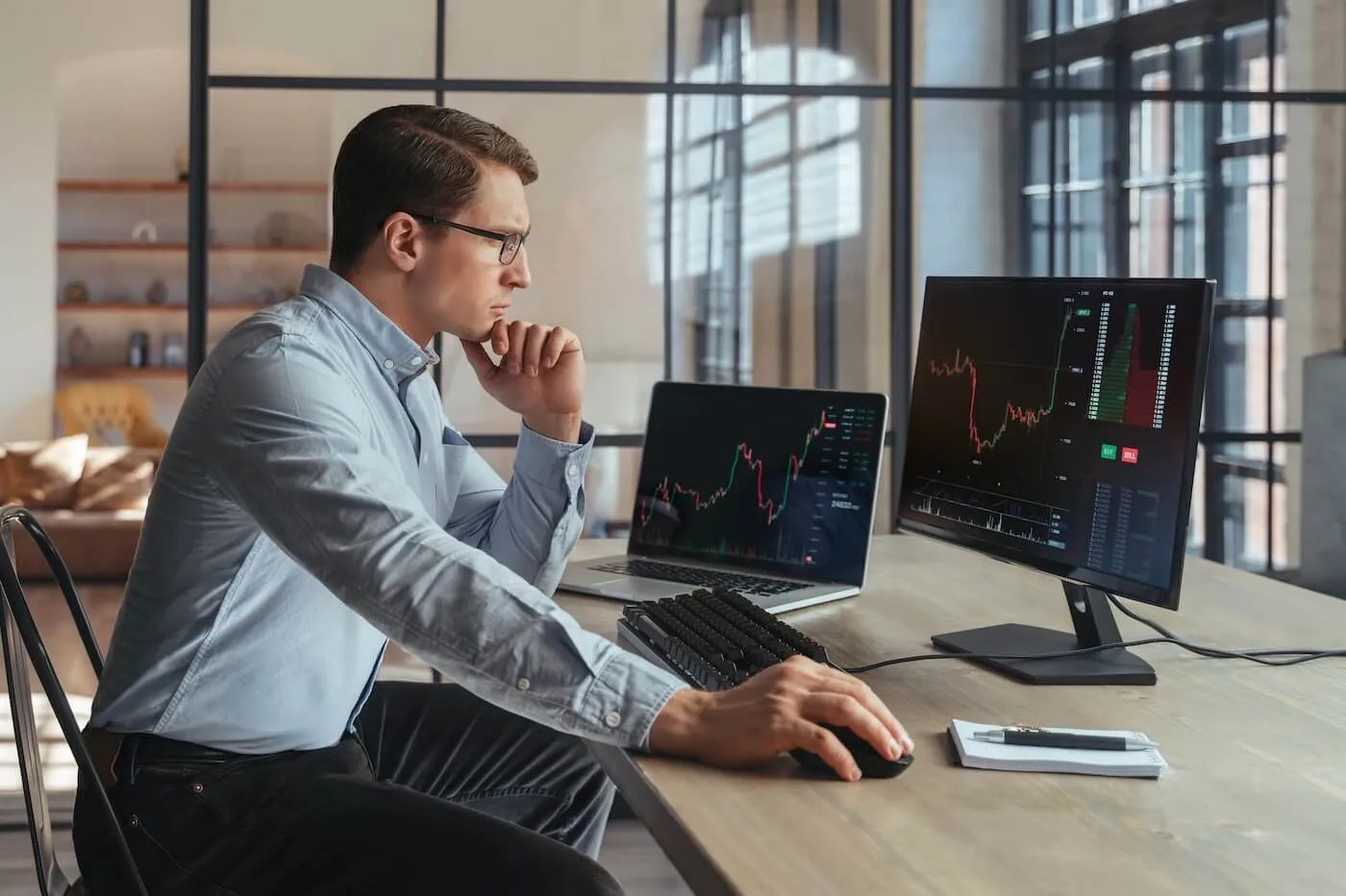 Focused businessman sits at a desk using a computer and laptop to monitor stock market charts on multiple screens in a modern office.