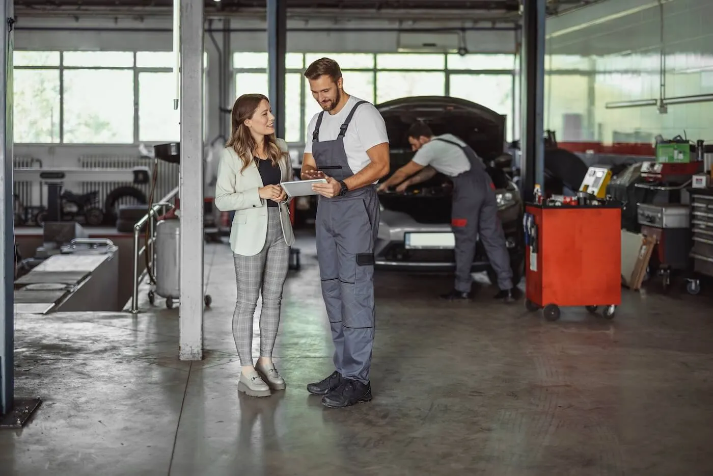 Male technician discussing a repair estimate with a smiling businesswoman in an auto shop, with another technician working on a car in the background.