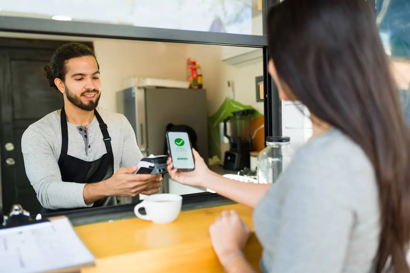 A woman at a café counter holds up a smartphone for contactless payment while a barista uses a card reader, with a cup on the counter.