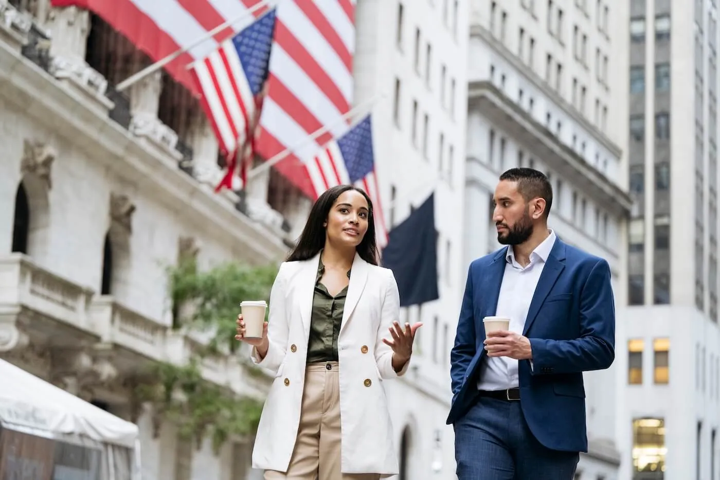 A man and a woman in business‑casual outfits walking down a city street holding coffee cups, with large American flags hanging from nearby buildings.