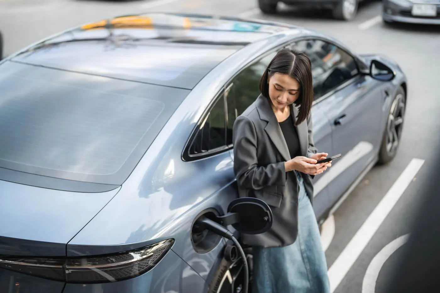 Young woman standing beside a parked electric car, holding a smartphone while the vehicle is connected to a charging cable, with other cars and a city street visible in the background.