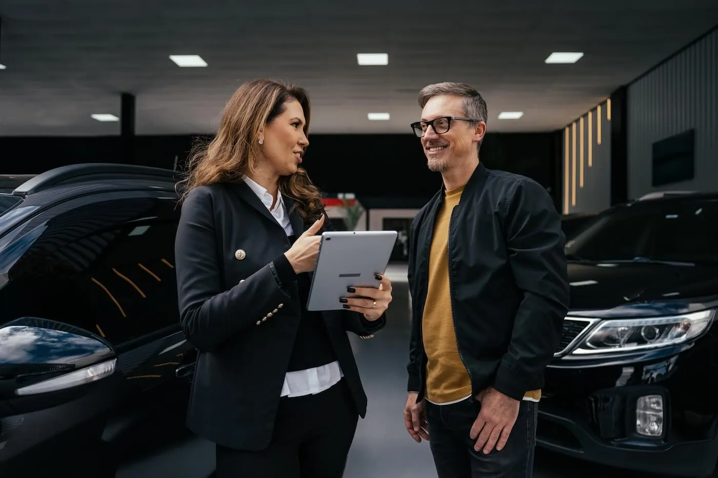 Smiling female consultant discussing auto loan options with a male customer, with black cars parked in the background