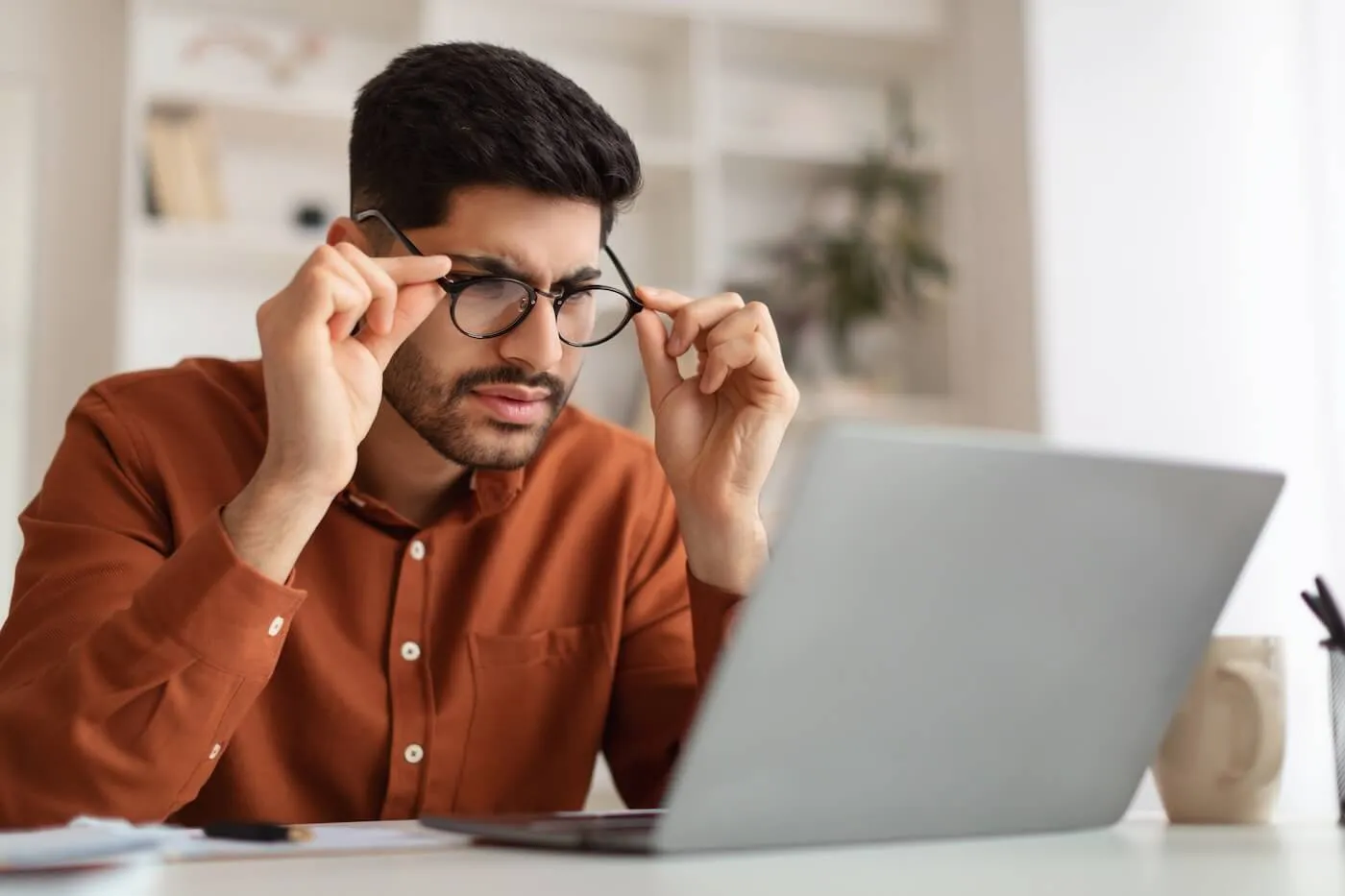 Concerned man seated at a desk looks closely at a laptop screen while adjusting eyeglasses with both hands, with a mug, papers, and office items on the desk and shelves visible in the softly lit background.