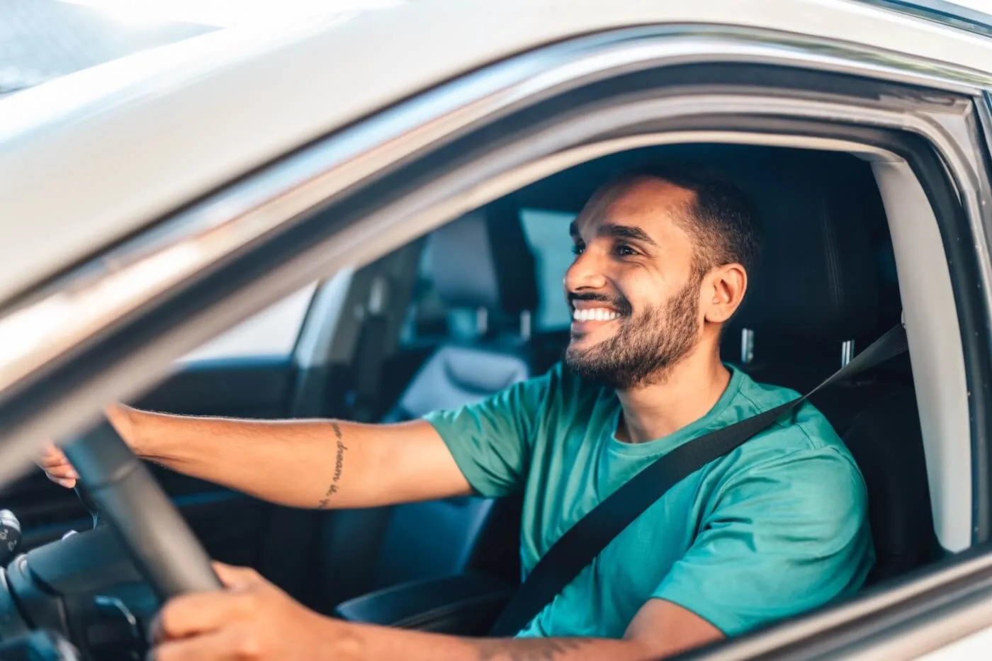 Smiling young man sits behind the steering wheel, wearing a seat belt, viewed through the open driver‑side window.
