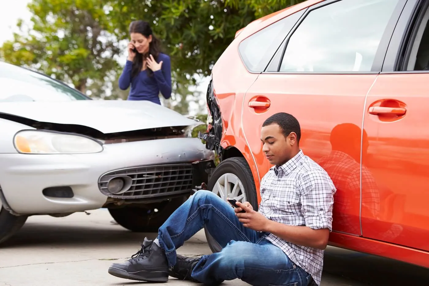 Two people near the scene of a car accident, with the man sitting on the ground holding a phone and the woman standing beside the damaged vehicles while making a call